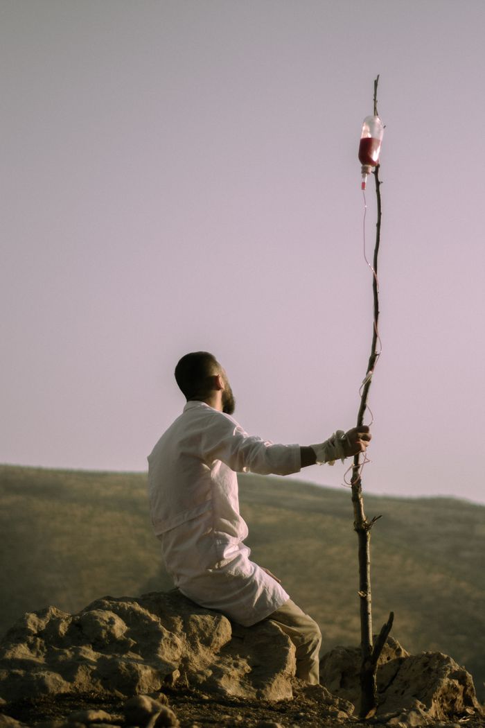 about-01 A man sits outdoors holding a pole with a medical infusion bag attached, symbolizing solitude and survival.